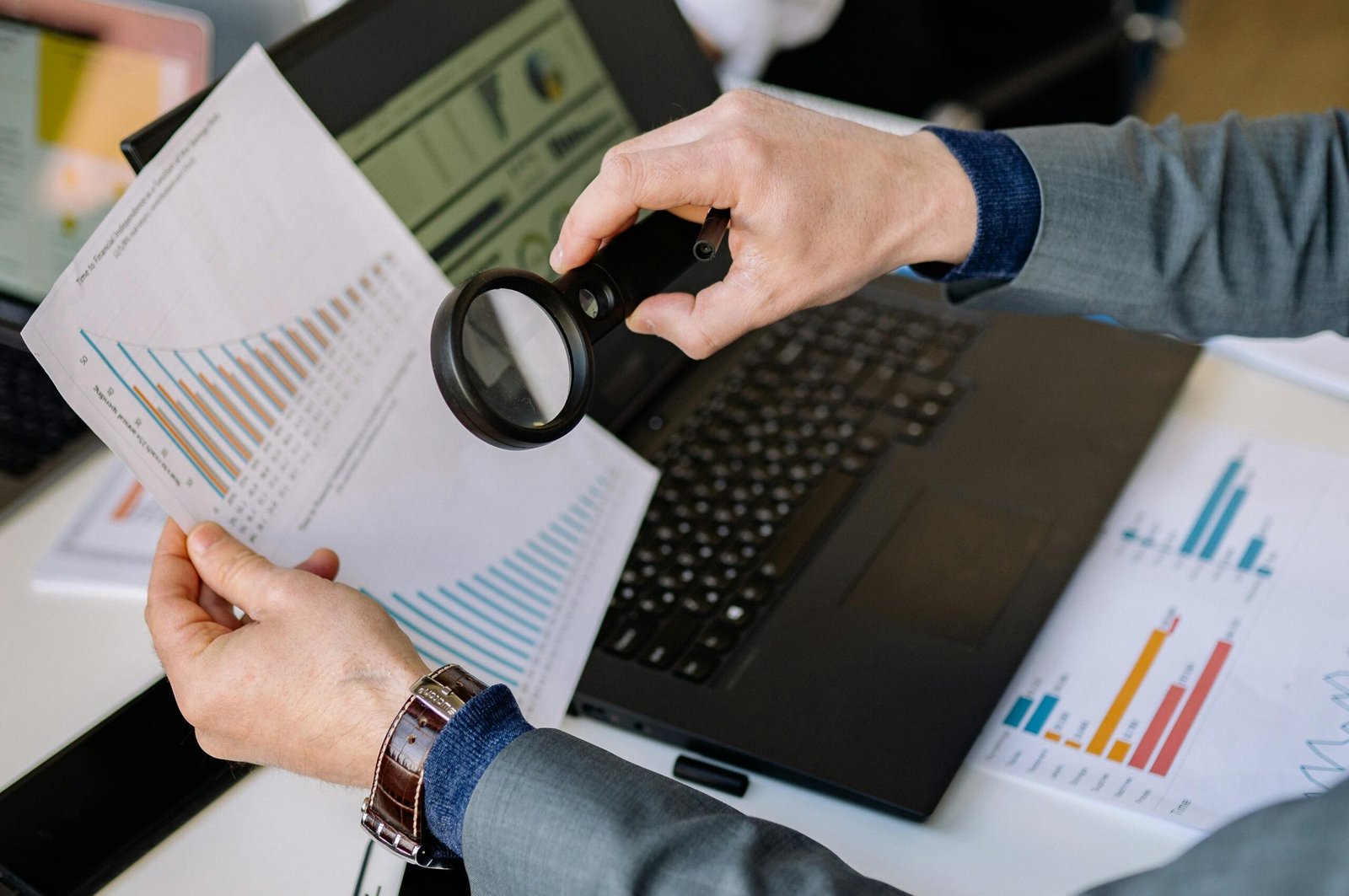 Businessman examines data charts with a magnifying glass over a laptop in an office setting.